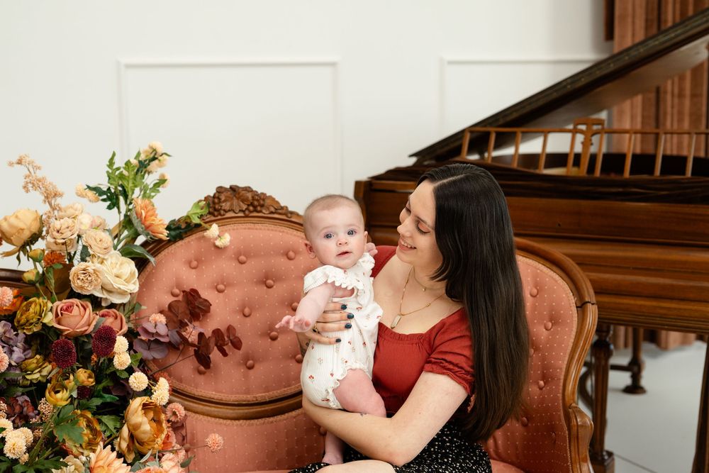 Mother in red top holds smiling baby on pink velvet chair beside floral arrangement near grand piano.