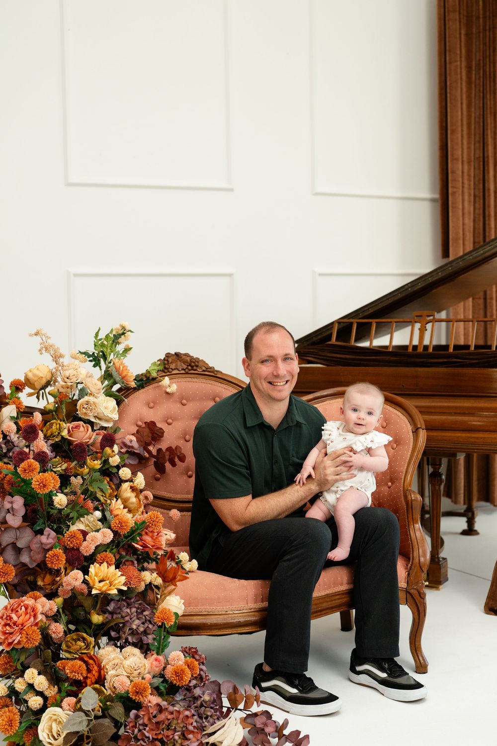 Father in green polo sits on pink velvet sofa holding baby girl beside colorful fall floral arrangement.