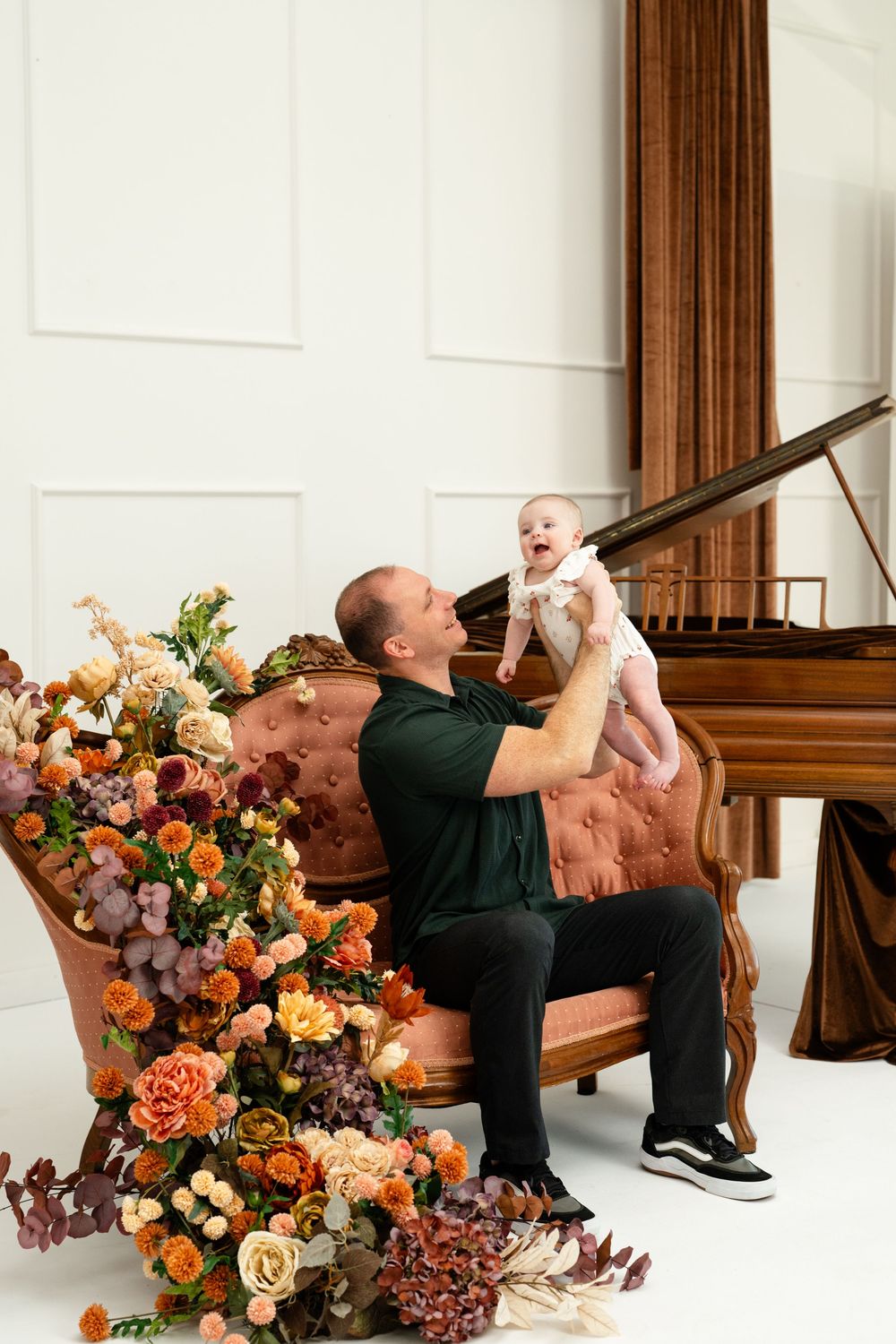 A father lifts his laughing baby on a pink velvet sofa surrounded by lush floral arrangements near a grand piano.