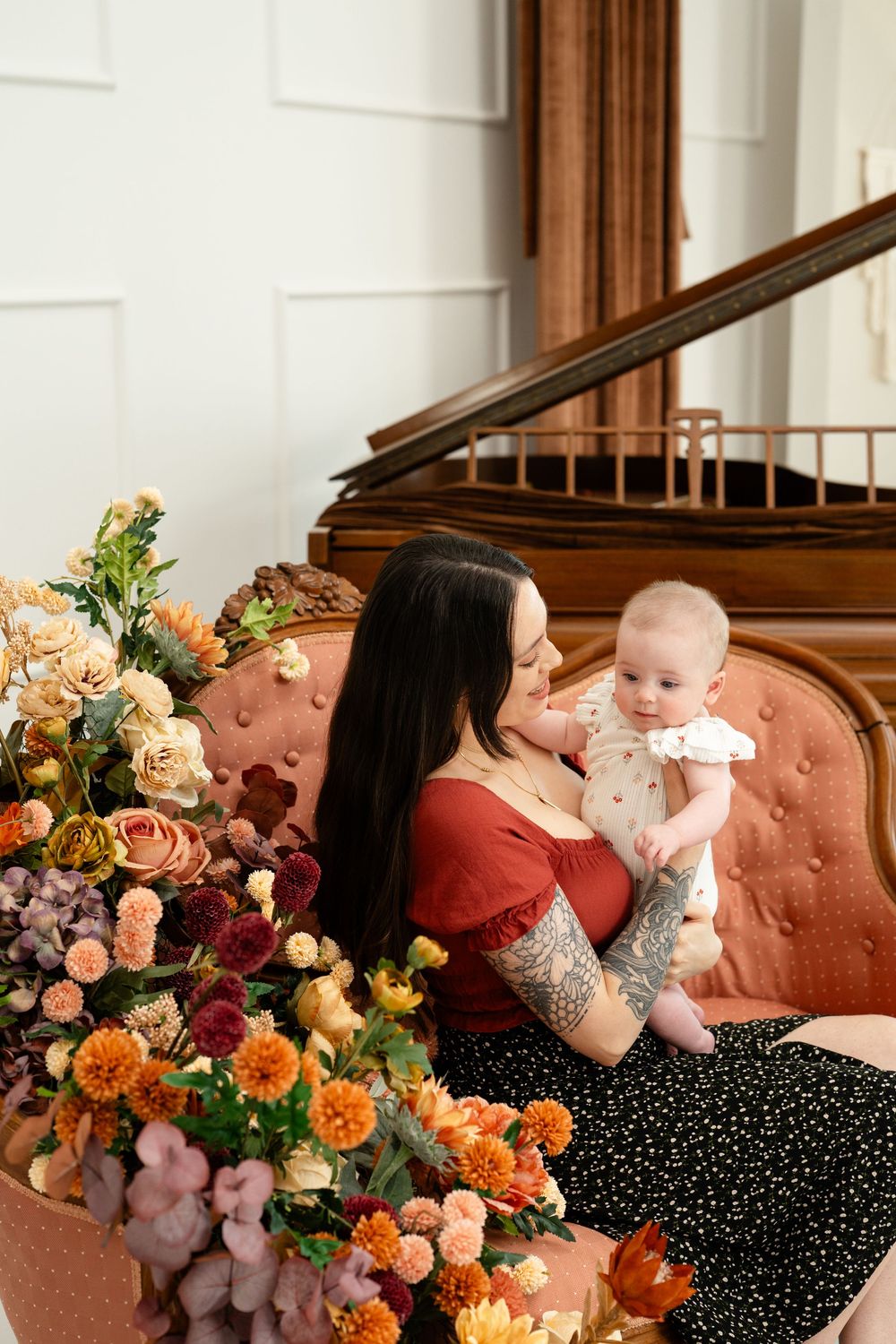 A tattooed mother in a red top holds her baby on a pink chair surrounded by colorful autumn flowers near a grand piano.