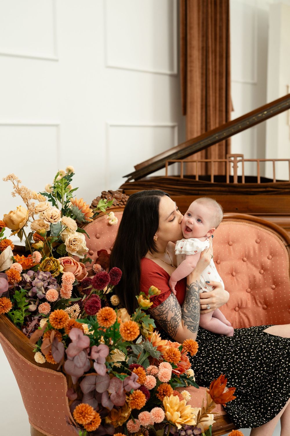 Mother kissing her baby while seated on a pink tufted sofa surrounded by autumn floral arrangements.
