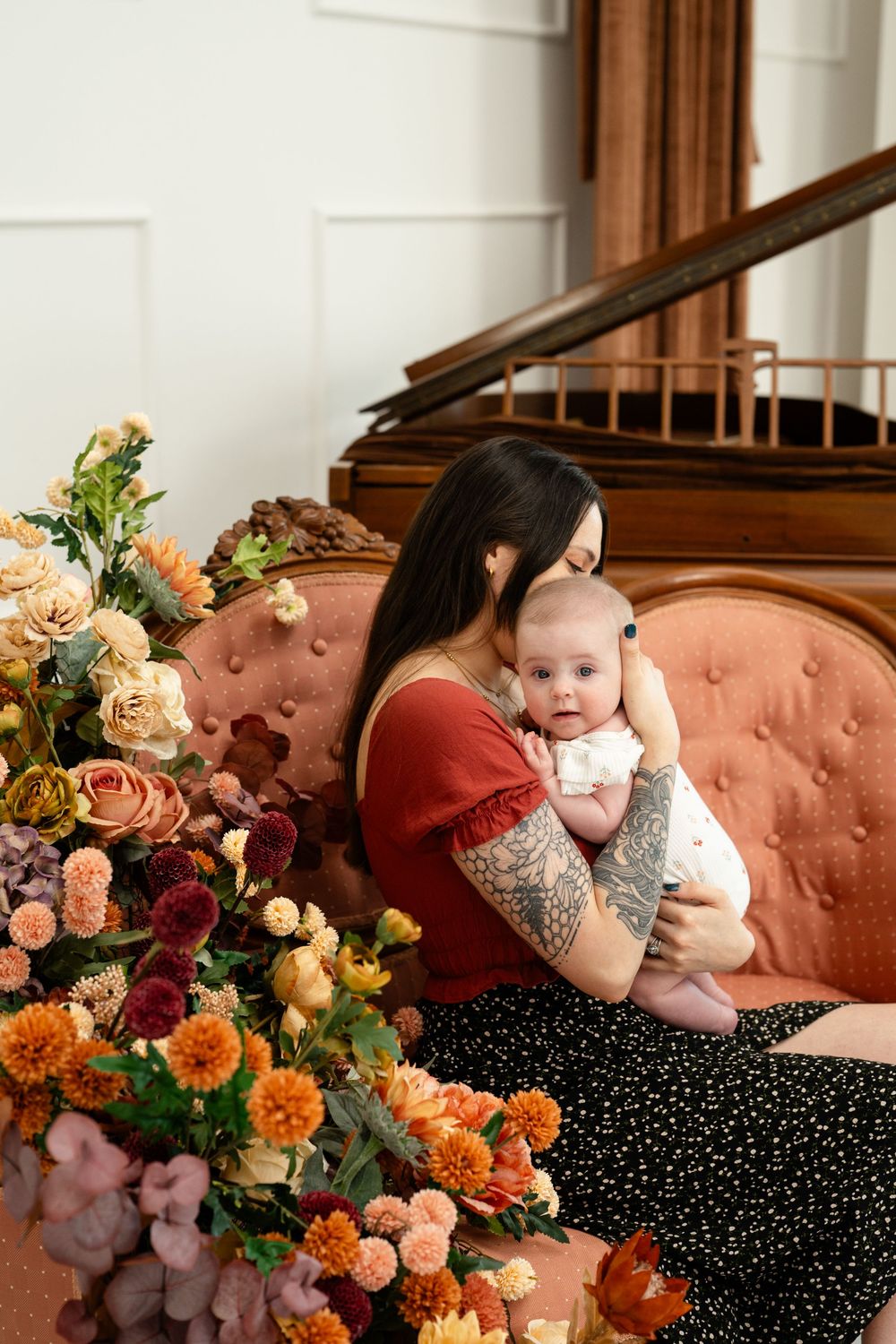 Mother tenderly holding her infant on a pink velvet sofa beside vibrant fall flower bouquets.