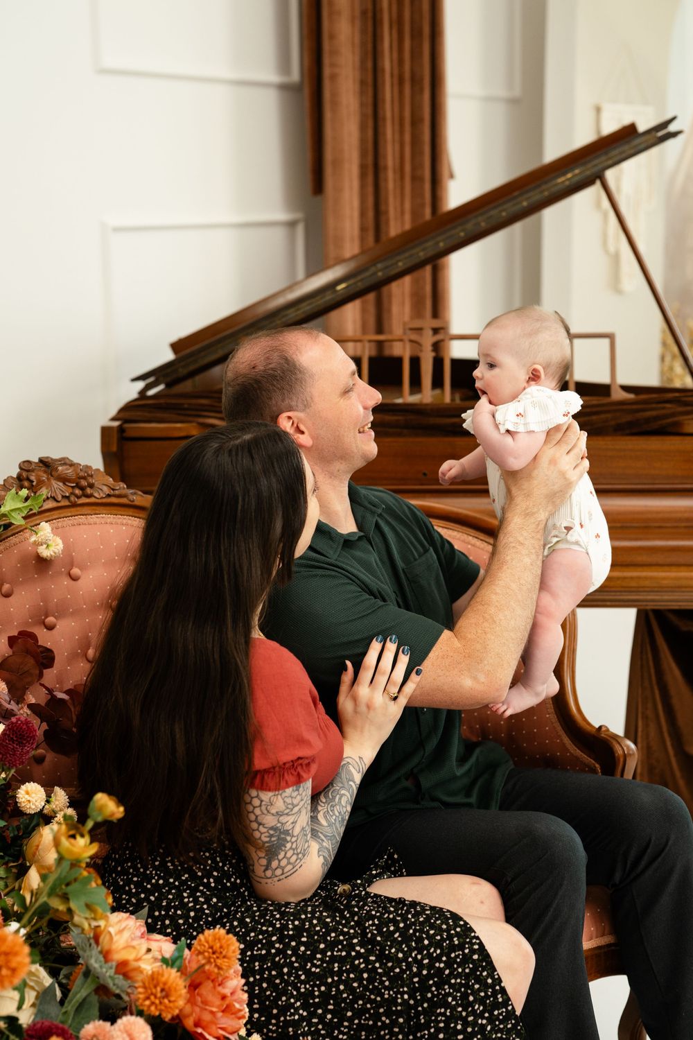 A couple sits by a grand piano holding their newborn baby, surrounded by autumn flowers in a warm studio setting.