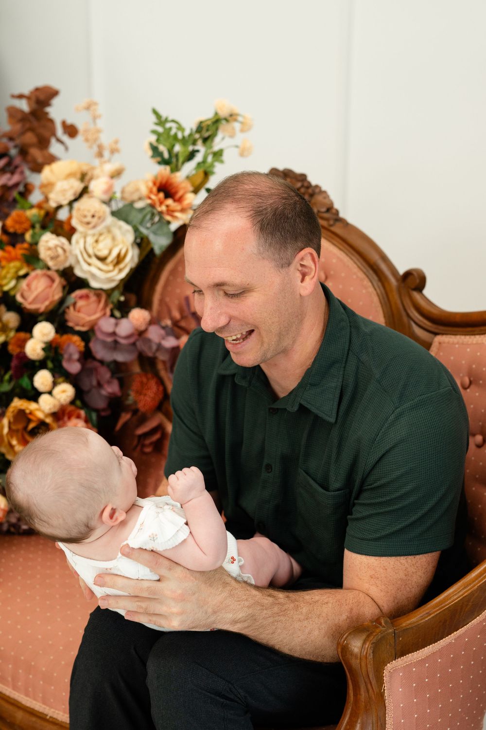 Smiling father holding newborn baby in a chair with autumn floral arrangement in a portrait studio setting.