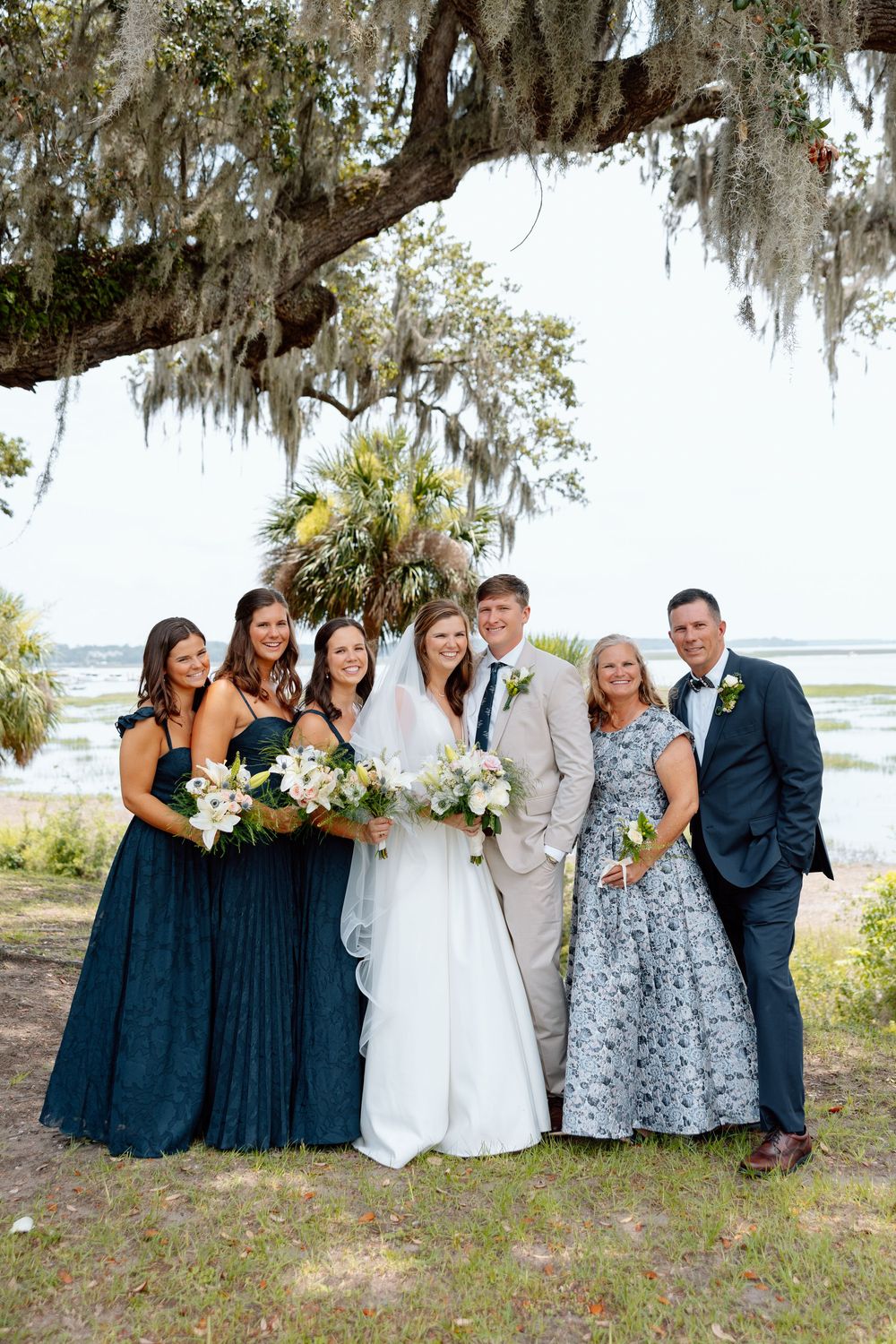Wedding party poses under Spanish moss tree by waterfront, bridesmaids in teal gowns with bride and groom.
