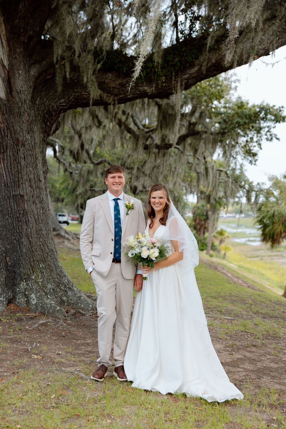 Bride and groom pose under a moss-draped oak tree, she holds a white bouquet, he wears a tan suit.