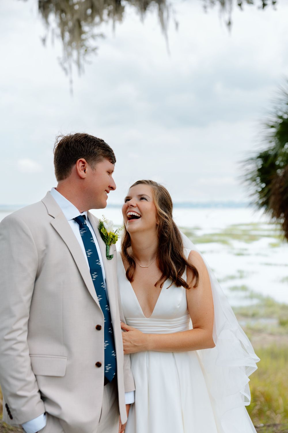 Smiling bride and groom share a joyful moment outdoors near a waterfront, groom in tan suit with blue tie.