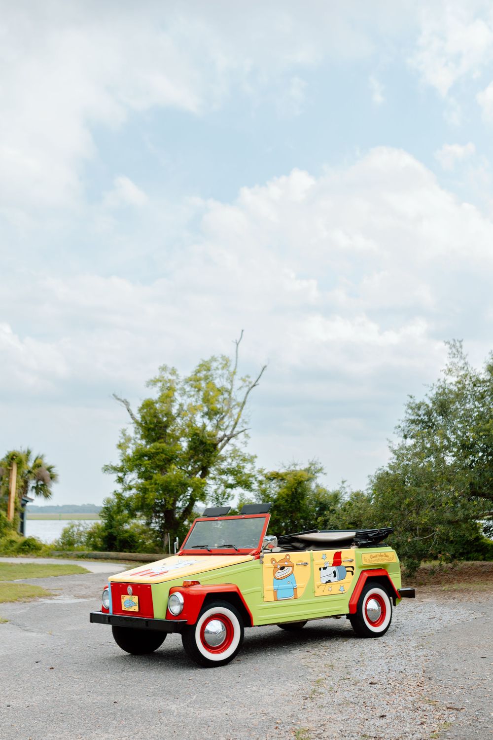 Colorful vintage VW Thing convertible with red and yellow paint and cartoon decals parked on gravel near waterfront trees.