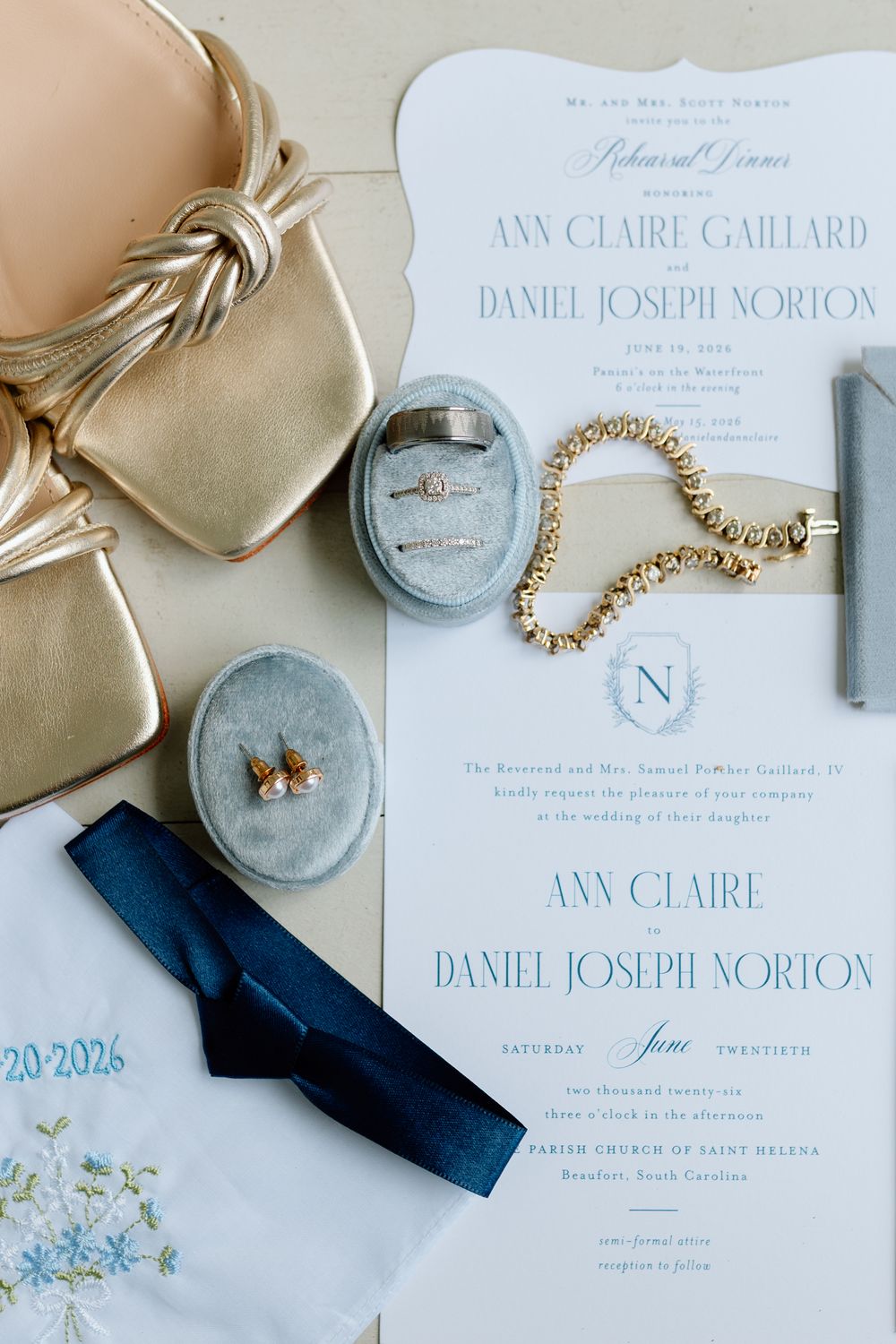Elegant wedding flat lay featuring rehearsal dinner invitation, gold heels, jewelry, navy tie, and velvet ring box on soft blue background.