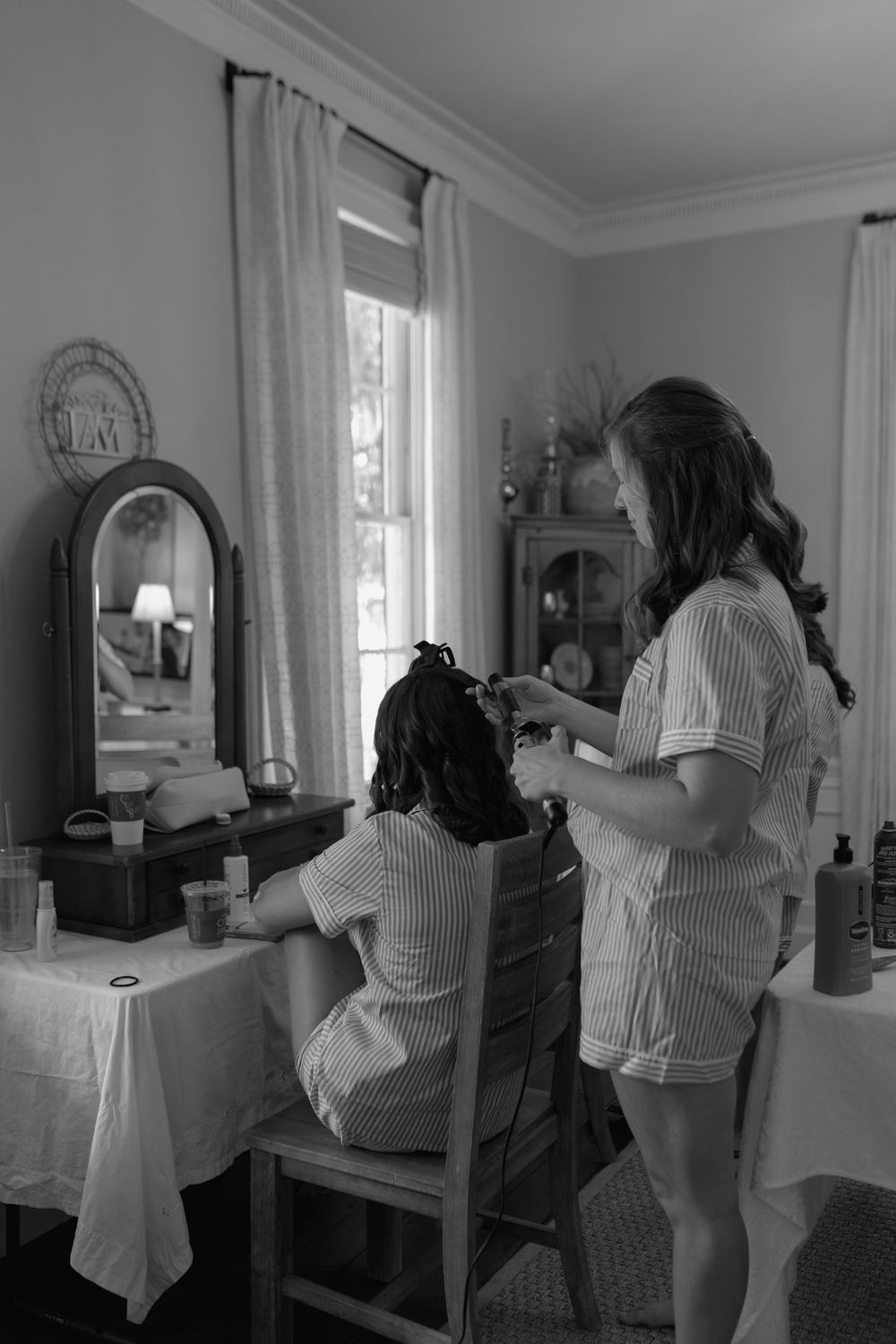 Black and white photo of a woman styling another woman's hair at a vanity mirror in an elegant room.