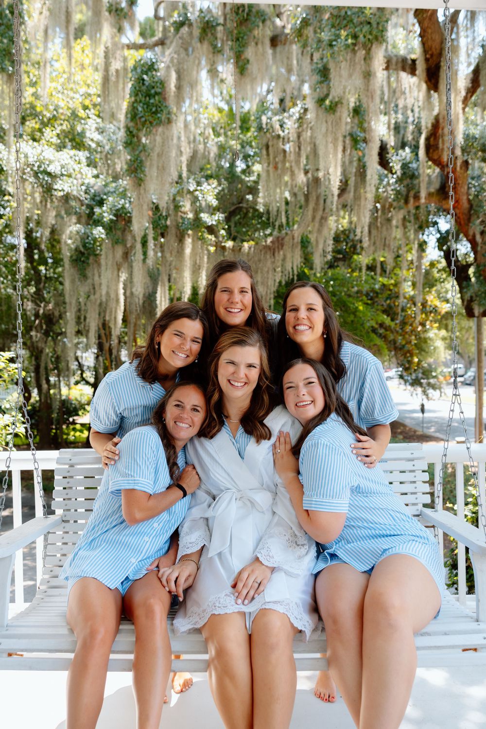 Bride in white robe with bridesmaids in blue seersucker on porch swing under Spanish moss.
