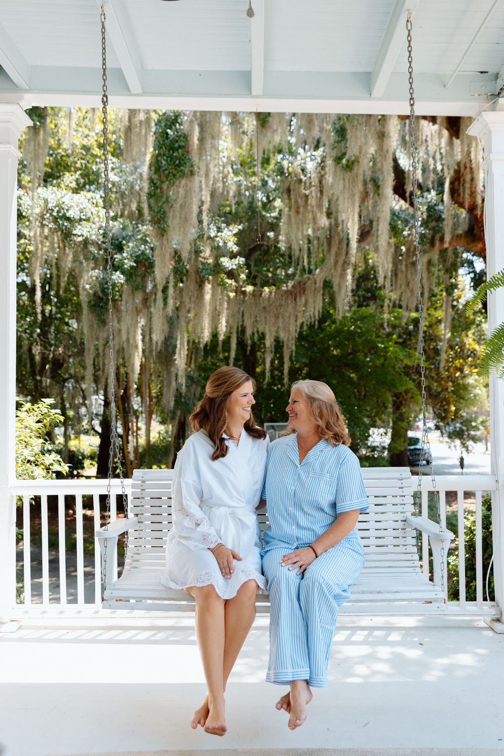 Two women in pastel pajamas sit on a white porch swing surrounded by Spanish moss-draped trees.