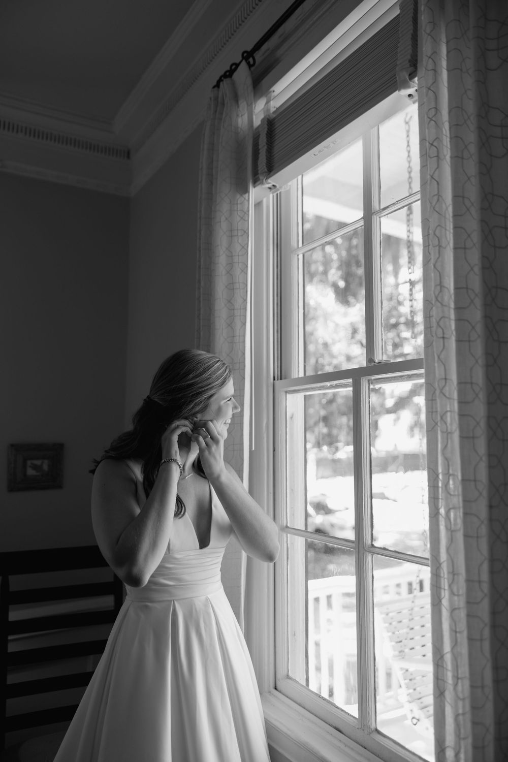 Black and white photo of a bride in a white gown adjusting her earring while standing by a sunlit window.