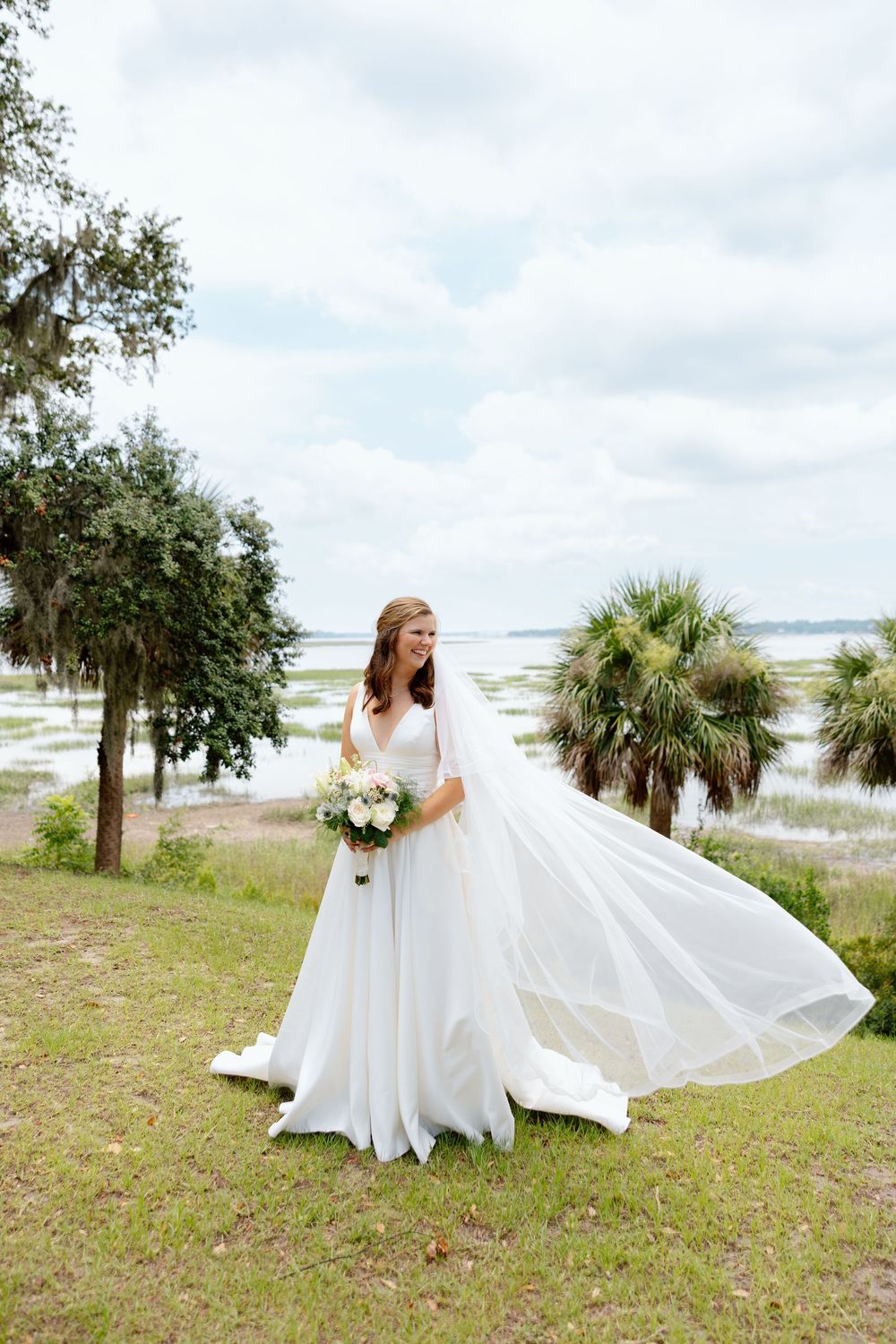Bride in flowing white gown with veil blowing in wind holds bouquet near waterfront with palm trees.