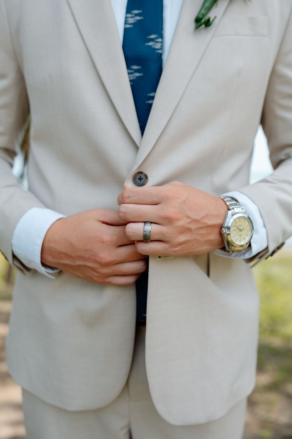Groom in beige suit with blue tie, wearing a silver wedding band and watch, buttoning his jacket.