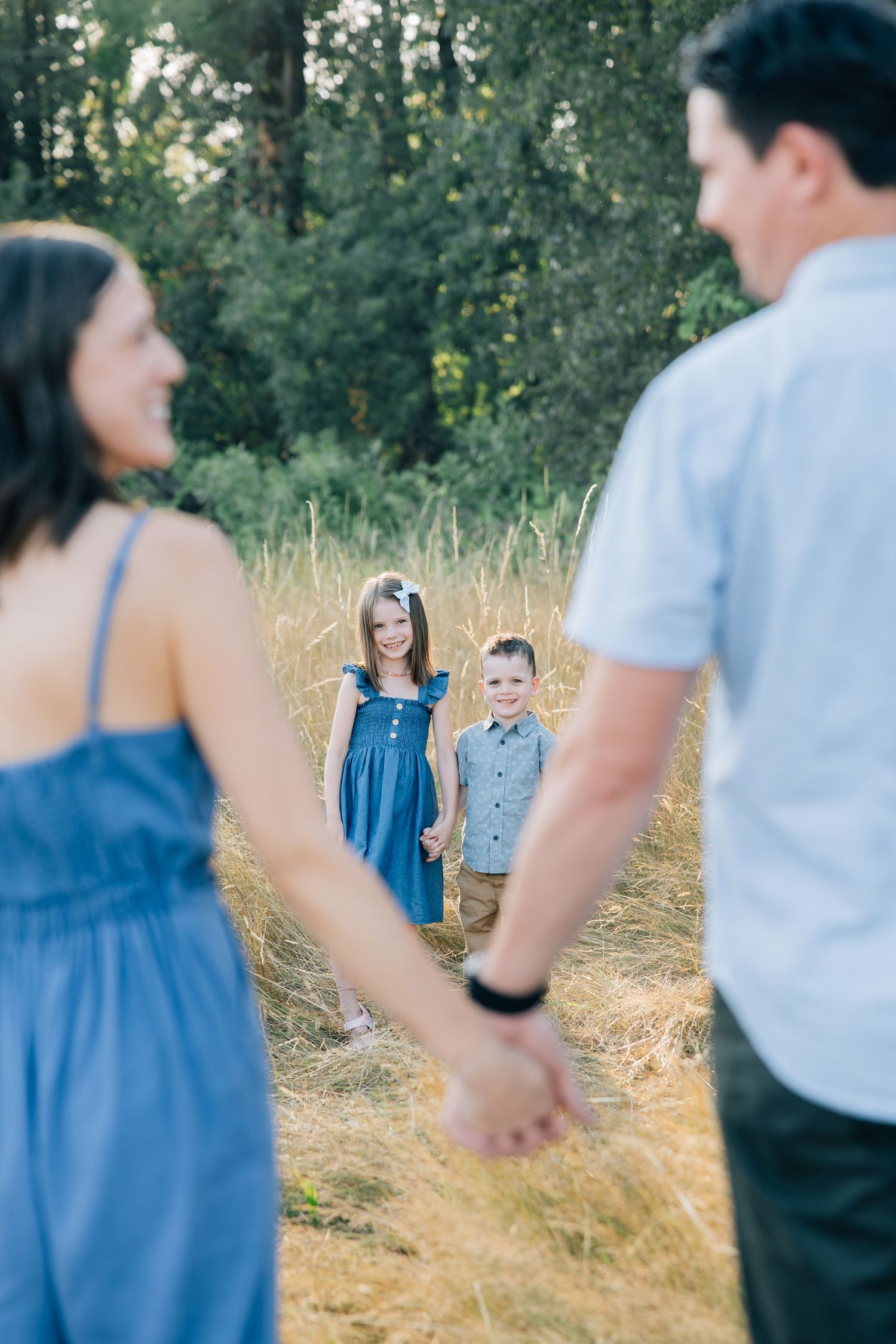Family holding hands in golden field, smiling children in blue outfits with tall trees in background.
