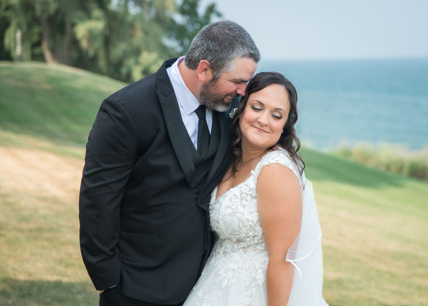 Bride and groom share a tender moment outdoors near the ocean, with the groom in a black suit nuzzling the smiling bride.