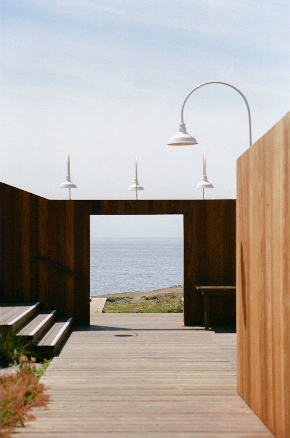 Modern wooden deck with gooseneck lights frames ocean view through rustic corten steel walls under clear sky.