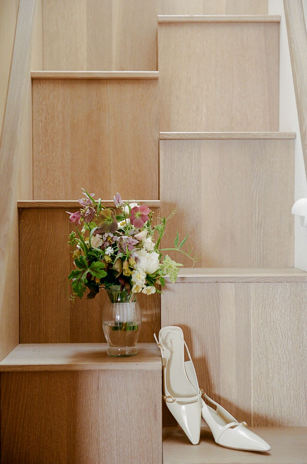 White bridal heels and a floral bouquet in a glass vase displayed on light oak wooden stairs.