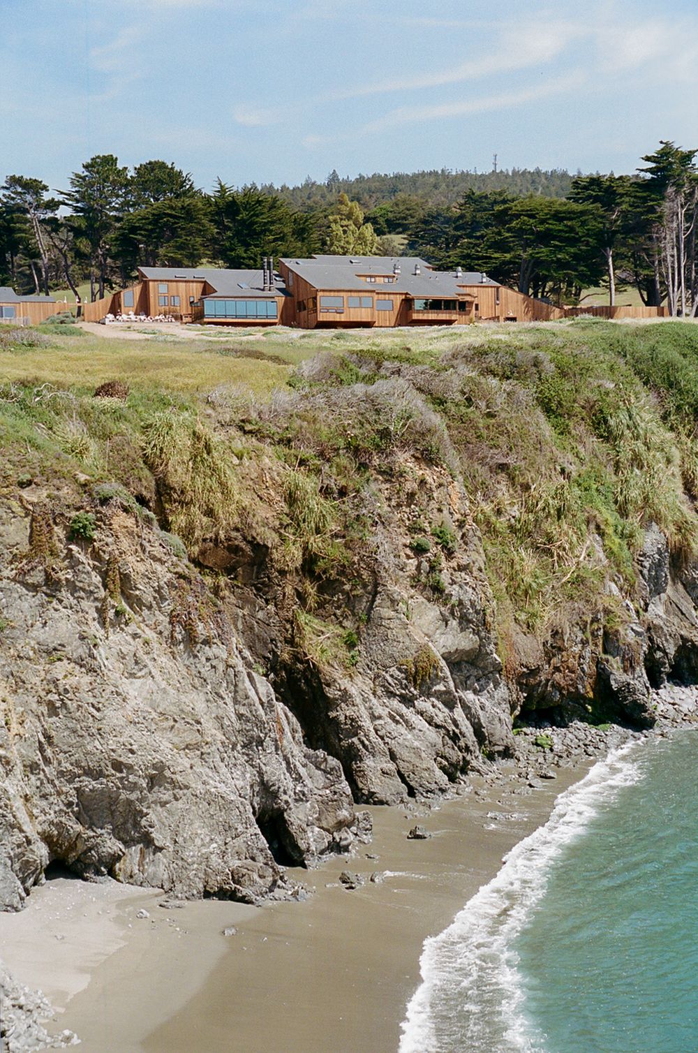 Wooden coastal homes perched on dramatic cliffs overlooking a sandy beach with turquoise waves and lush trees.