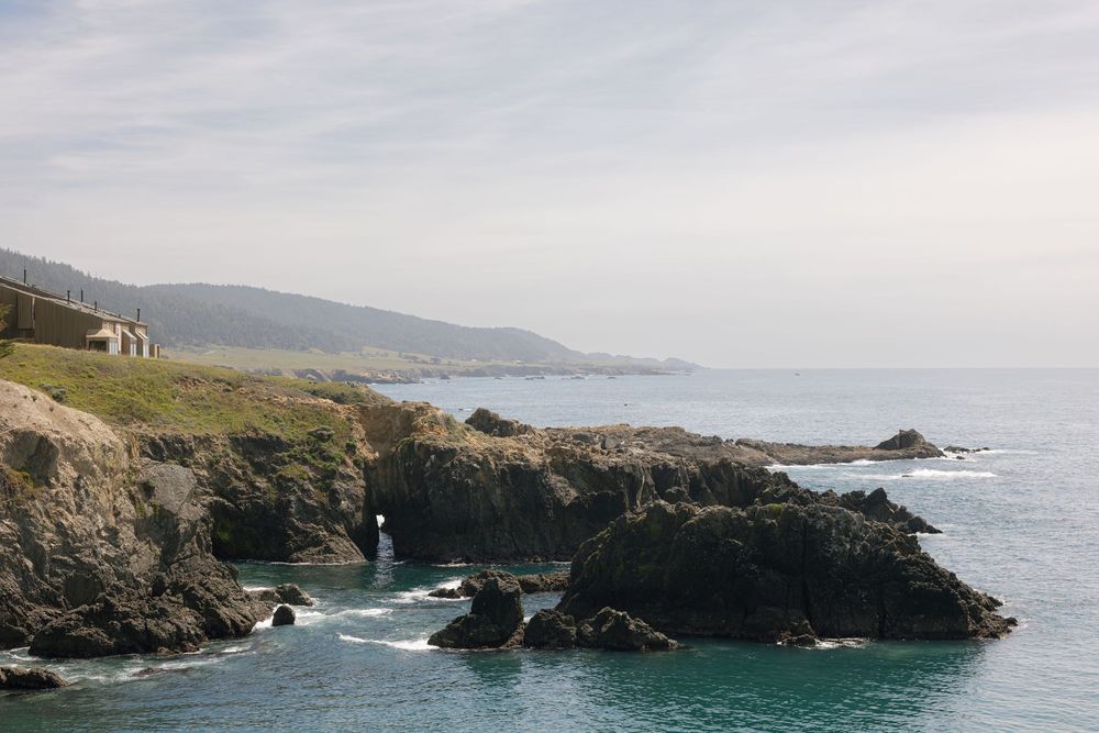 Rocky coastal cliffs overlooking turquoise ocean waters with a small building on the shoreline and green hills in the background.