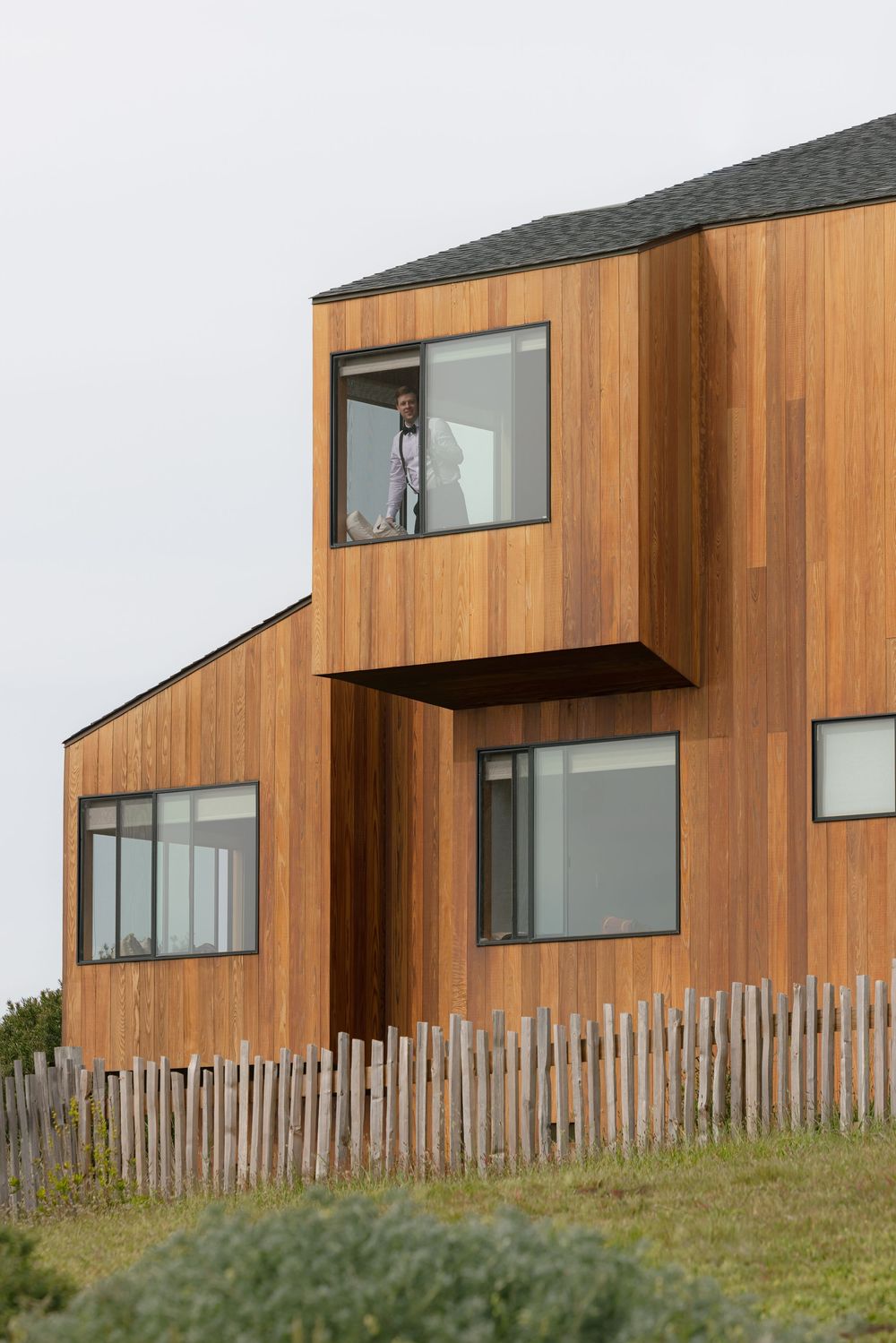 Modern cedar-clad two-story home with large windows and wooden fence, person visible on upper floor balcony.