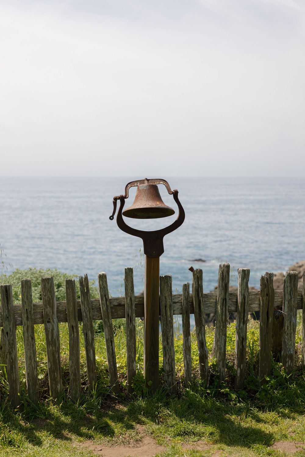 Rusty vintage bell on a post by a weathered wooden fence overlooking a calm blue ocean on a cloudy day.
