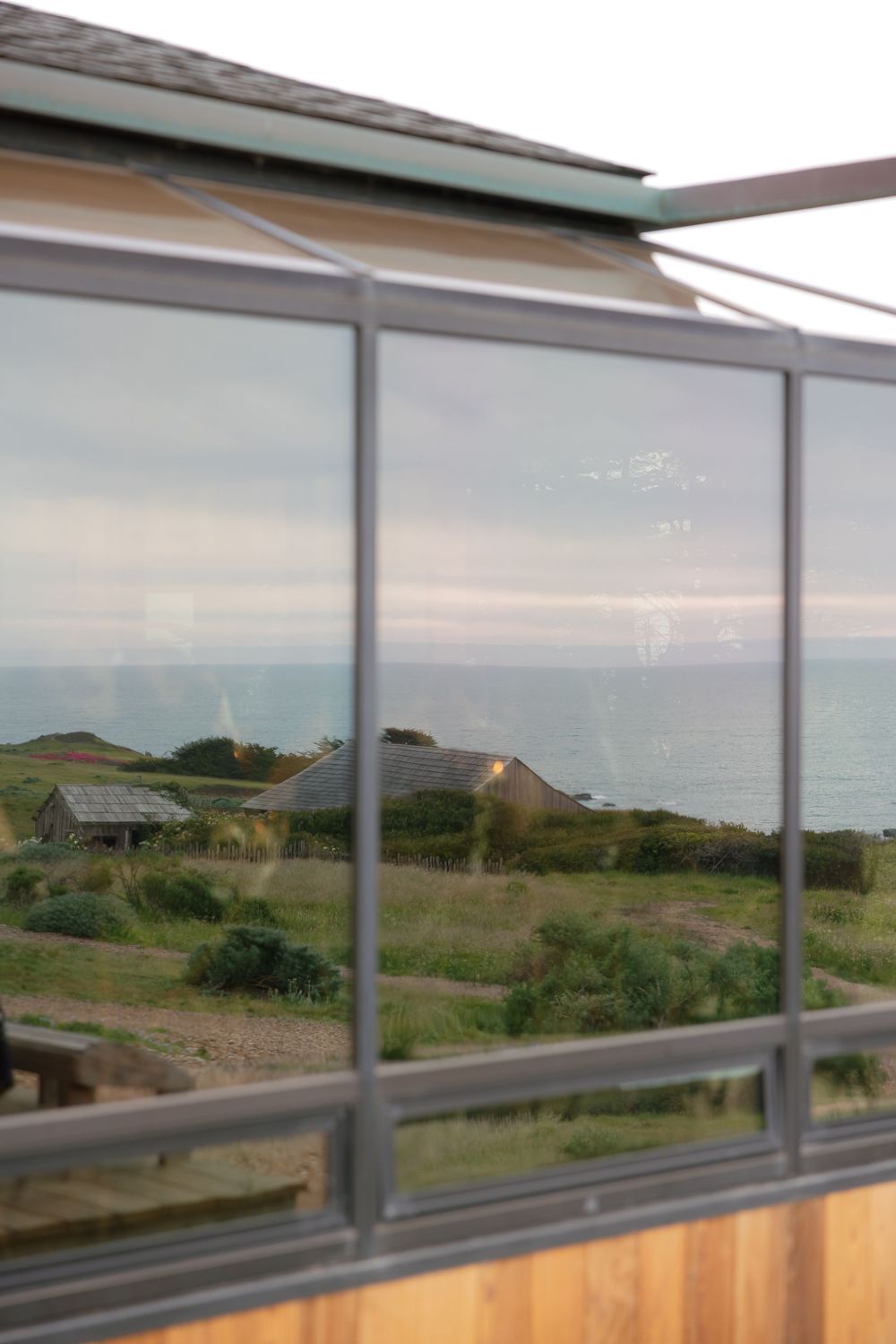 Reflective glass windows of a coastal building overlooking green fields, cottages, and a calm ocean under overcast skies.
