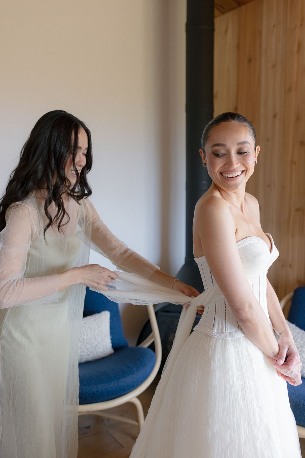 Bride smiling in strapless white wedding dress while bridesmaid helps adjust her gown in a bright room.