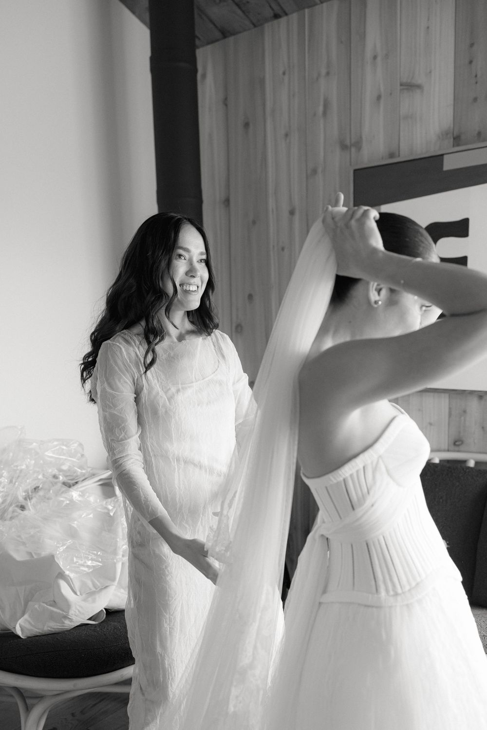 Black and white photo of a bride having her veil placed by a helper in a rustic wooden room.