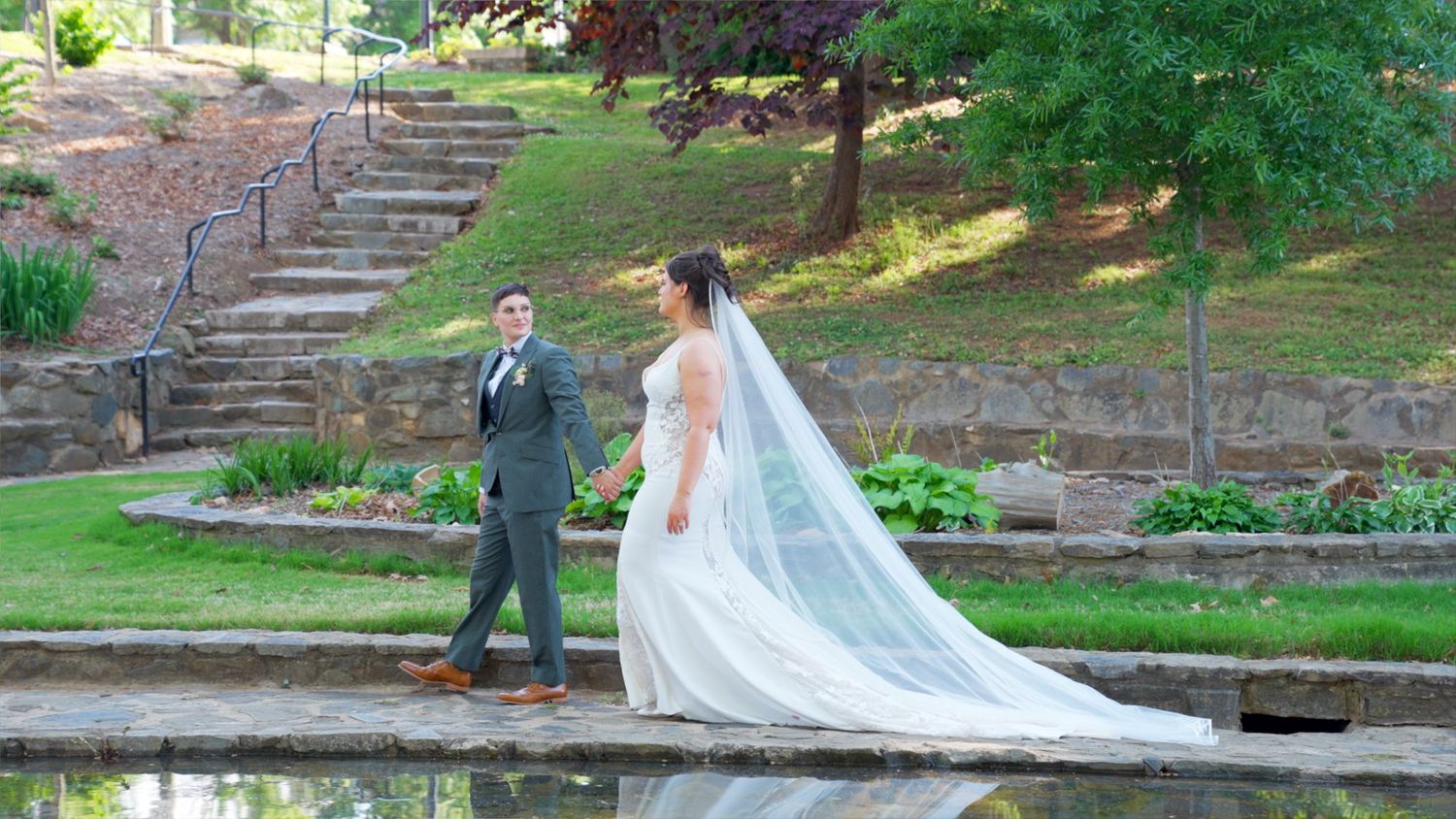 Two brides pose elegantly by a reflective pond with stone steps and lush greenery at an outdoor wedding venue.