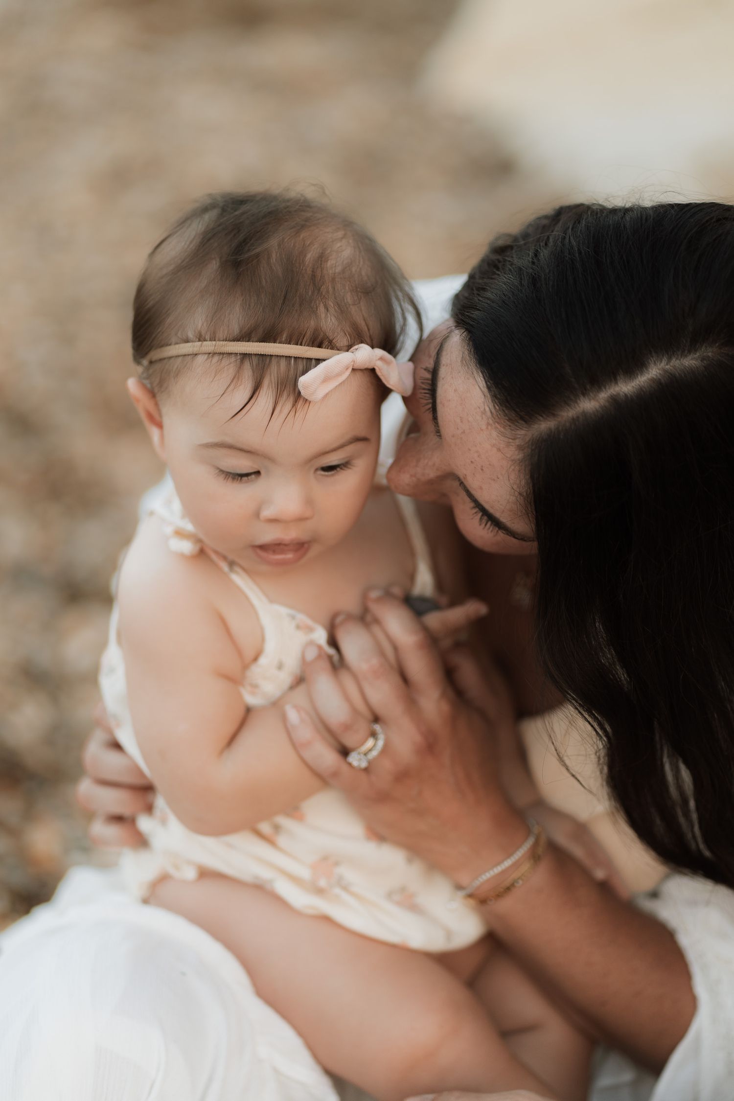A mother tenderly holds her baby girl wearing a pink bow headband and floral romper in a warm outdoor setting.