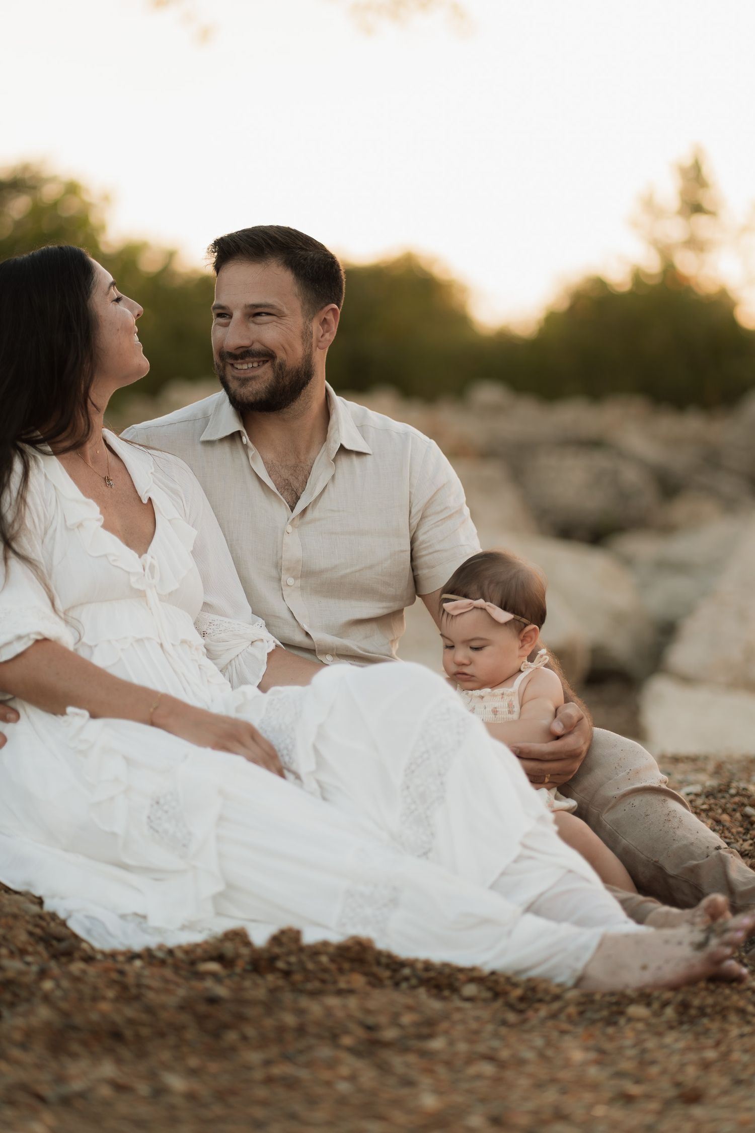 A happy family sitting outdoors at golden hour, with a smiling couple and their baby girl on a rocky beach.