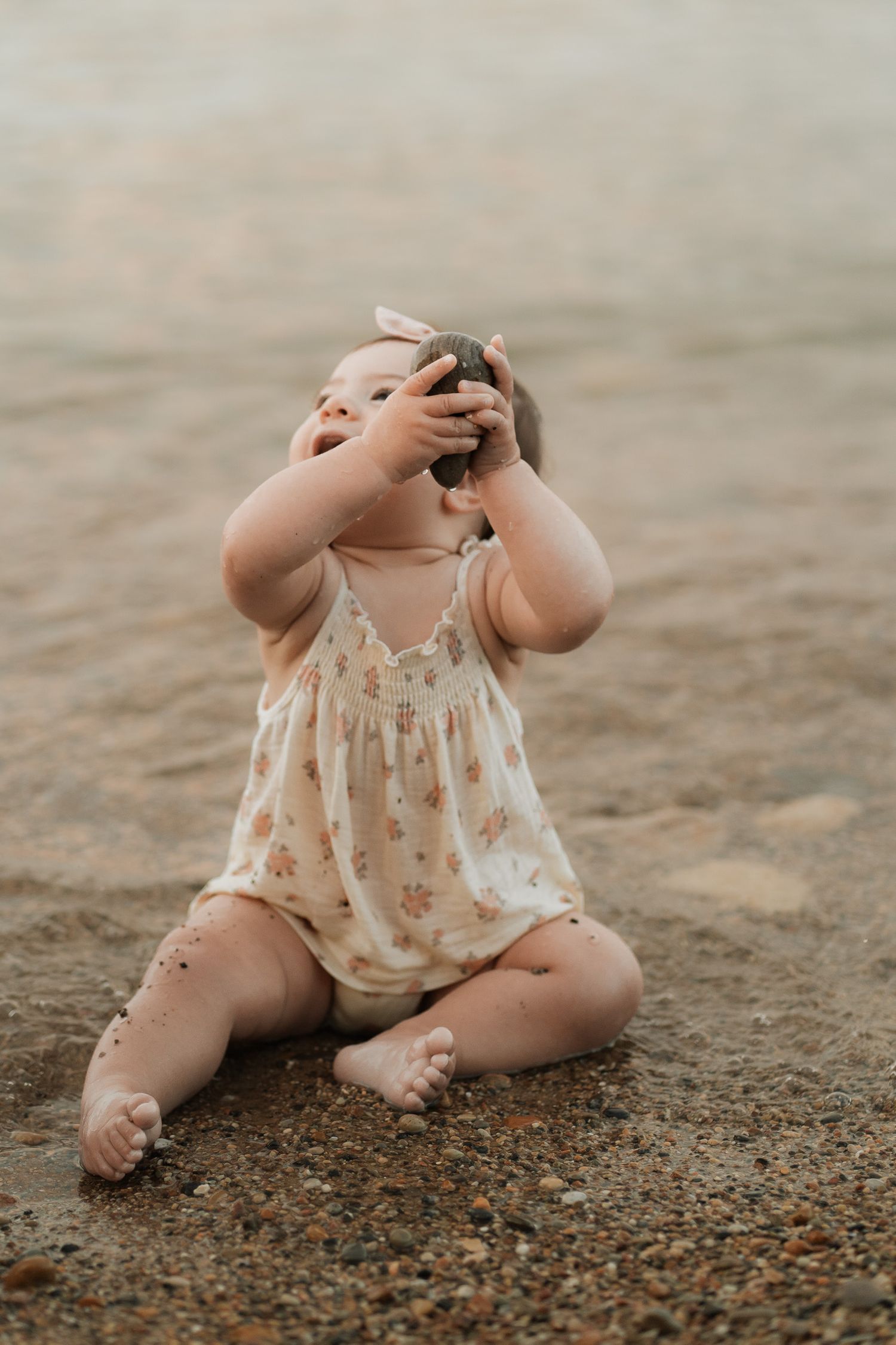 A baby girl in a floral romper sits on sandy beach, laughing while holding a rock up to her face.
