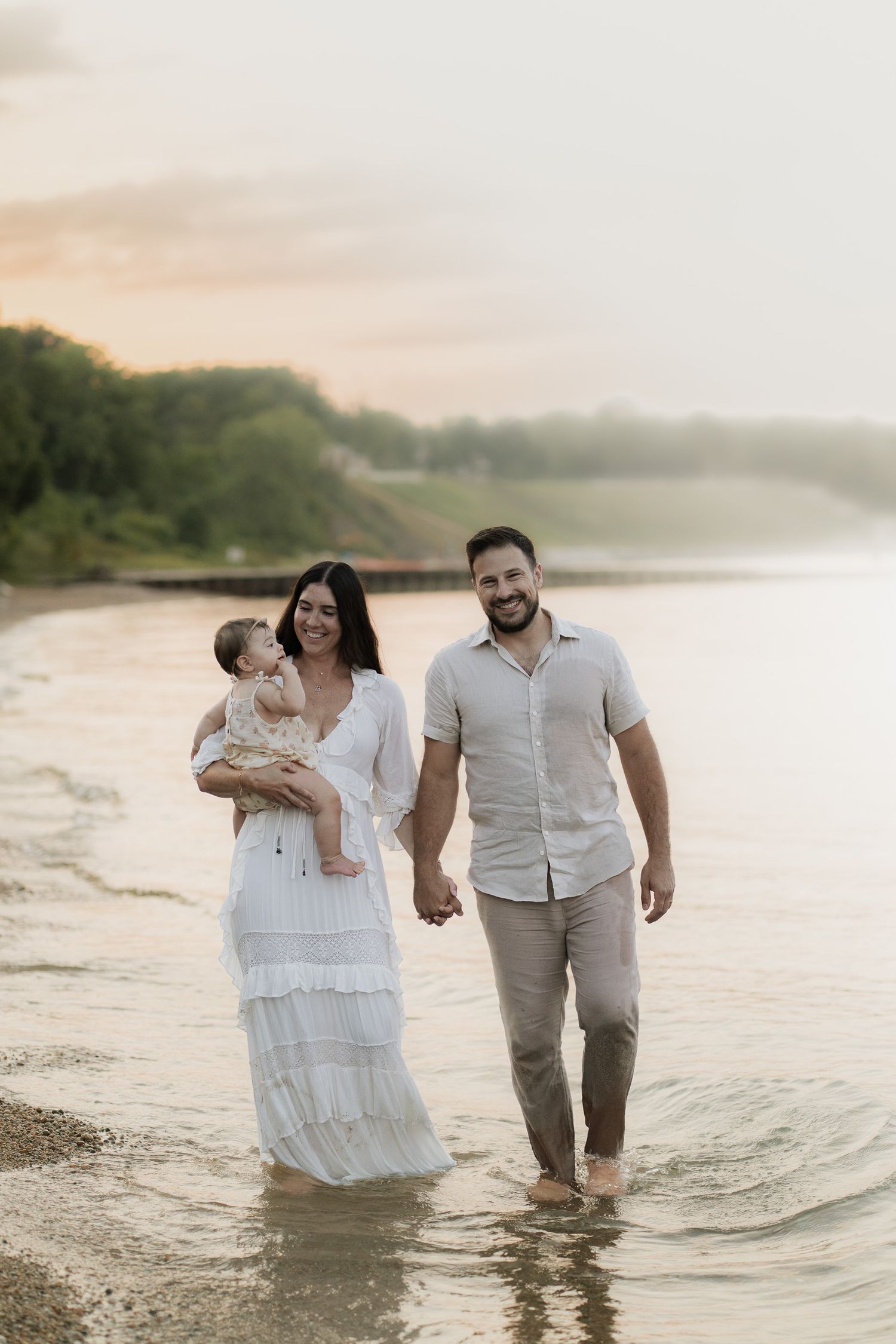 A young family walks along a sandy beach at sunset, with the mother in a white dress holding a baby.