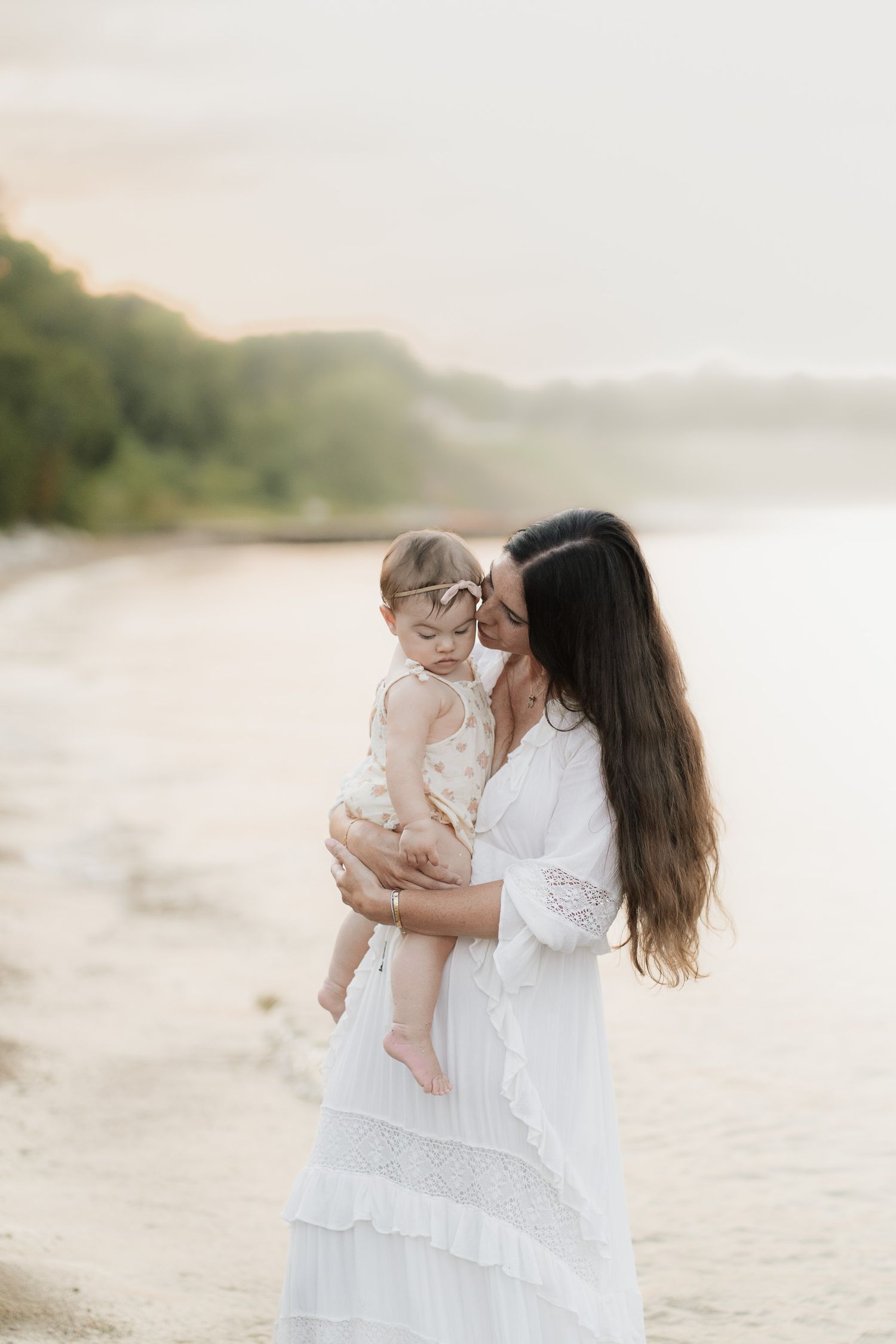 A mother in a white dress holds her baby girl on a serene beach at sunset, sharing a tender moment.