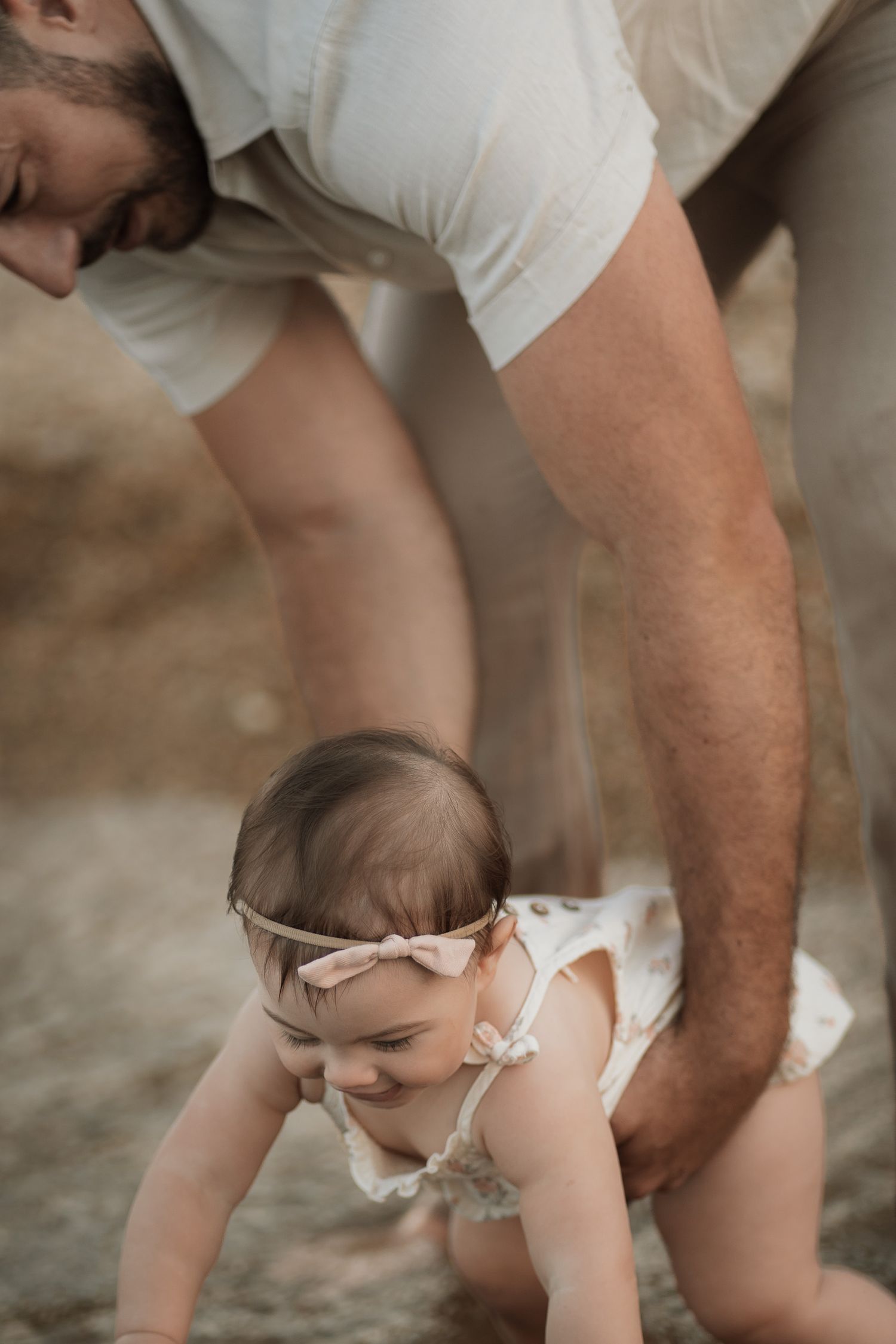 A father helps his smiling baby girl in a floral romper and pink bow headband learn to crawl outdoors.