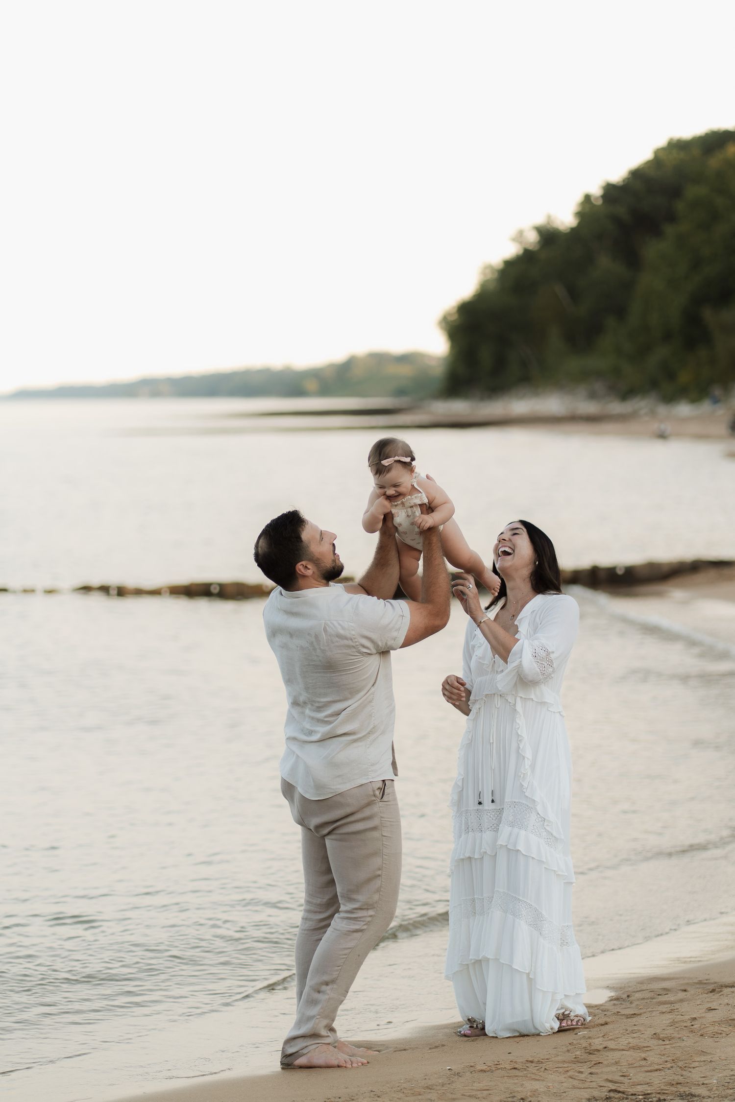 A happy family on a beach, with a father lifting a baby in the air as the mother laughs in a white dress.