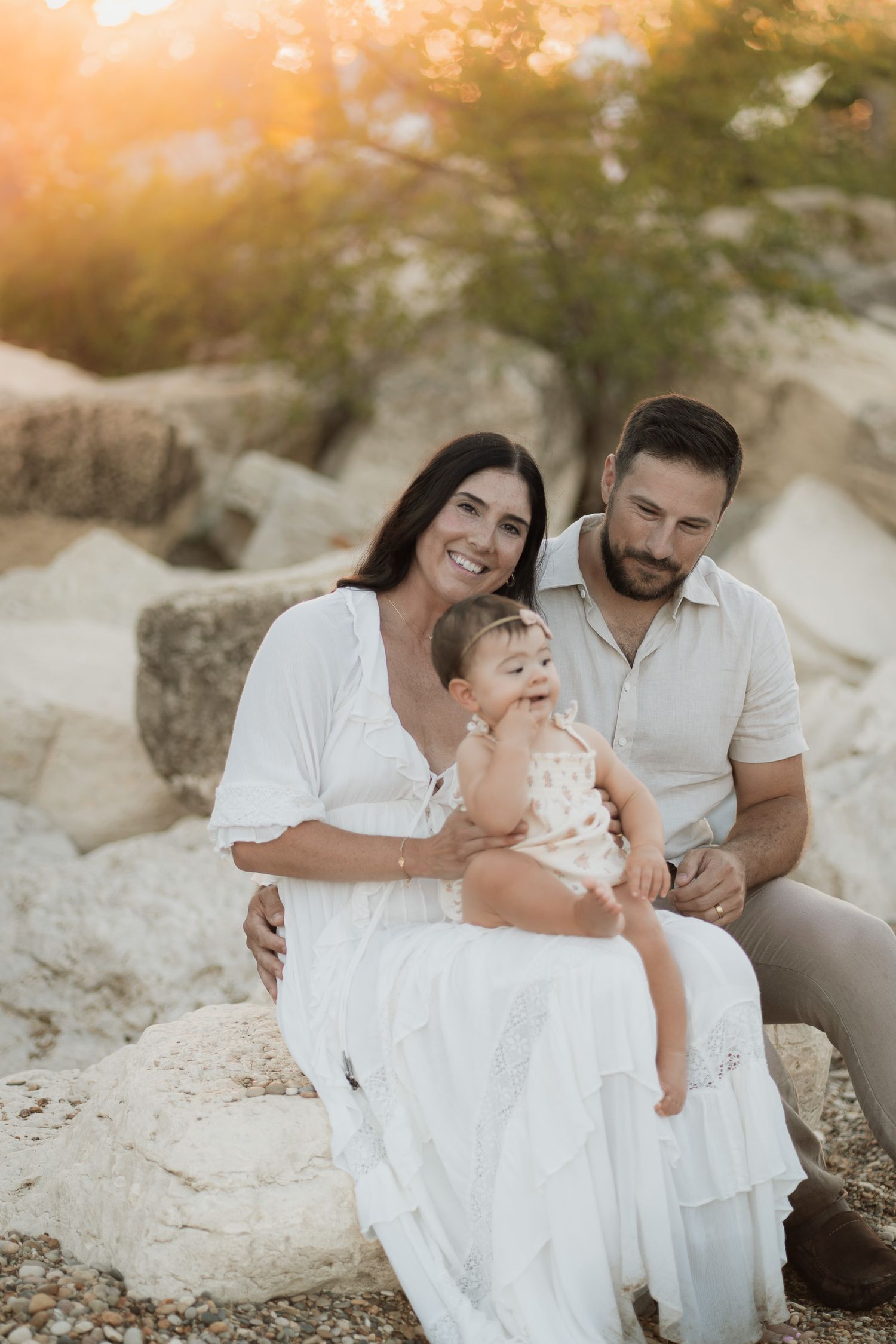 A smiling family of three sits on rocks at golden hour, parents in white outfits with their baby girl between them.