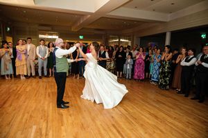 Bride and groom share their first dance on a wooden dance floor surrounded by wedding guests at a reception venue.
