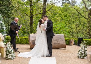 Bride and groom share first kiss at outdoor wedding ceremony with floral arrangements and officiant in wooded garden setting.