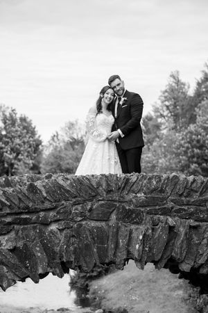 Black and white photo of a bride and groom smiling on a rustic stone bridge surrounded by trees.