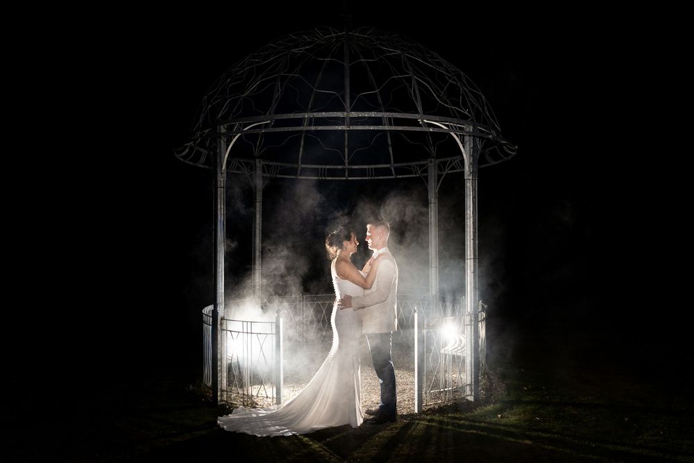 Bride and groom embrace under a glowing gazebo at night, surrounded by misty light in a romantic wedding portrait.