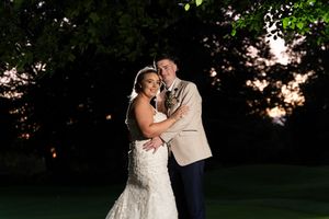 Bride and groom embrace outdoors at twilight, surrounded by lush trees with a warm sunset glow in the background.