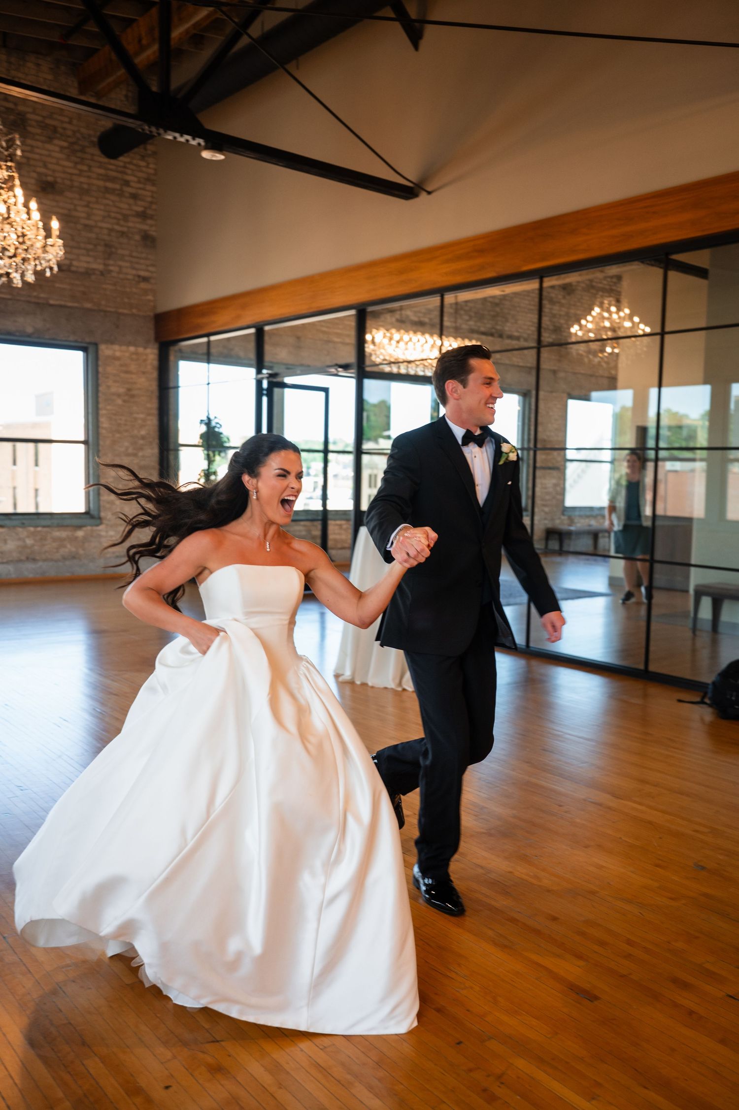 Joyful bride in white strapless gown and groom in black tuxedo making grand entrance at elegant wedding reception venue.