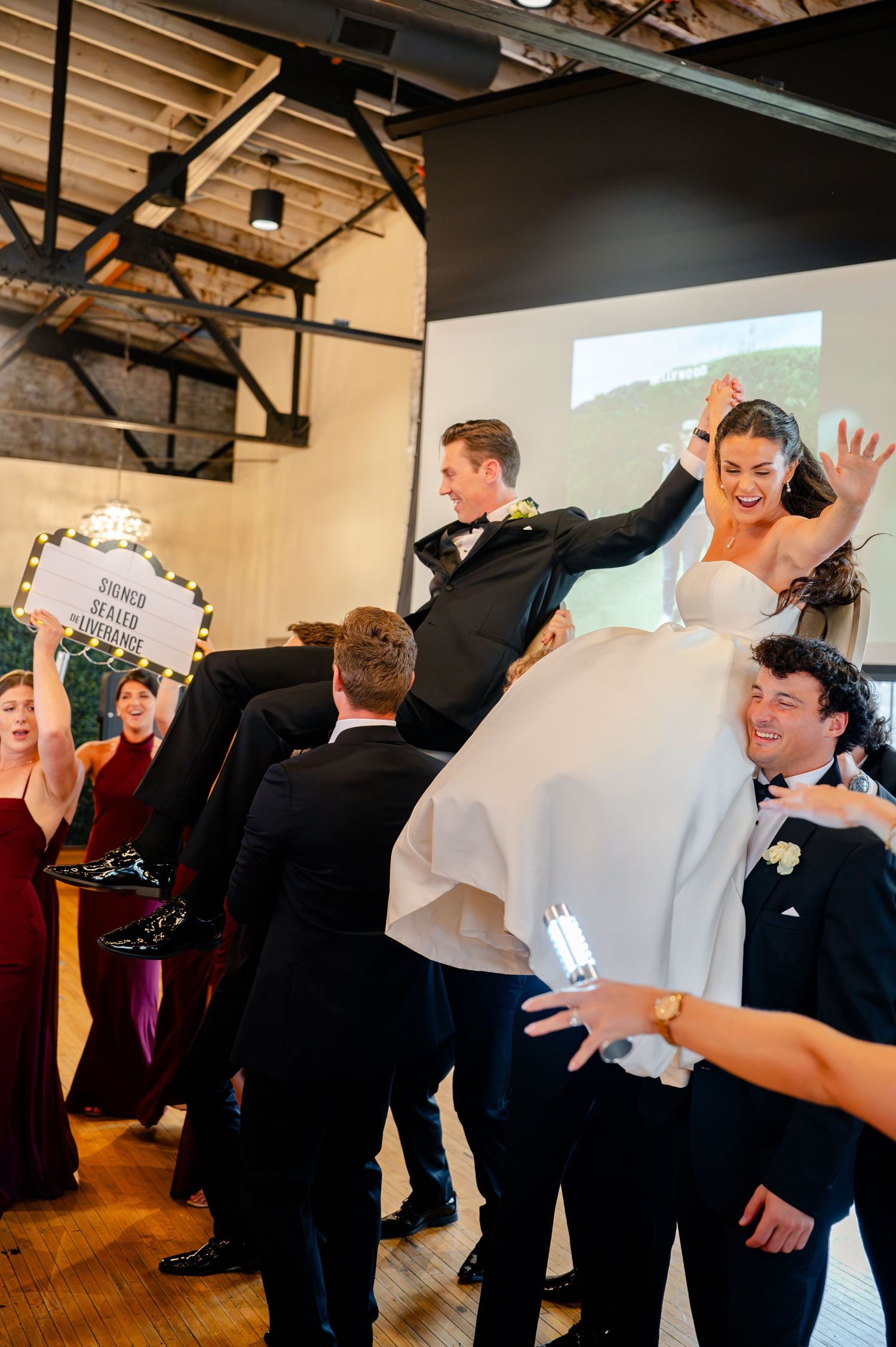 Bride and groom lifted by wedding guests during joyful reception celebration in modern industrial venue.