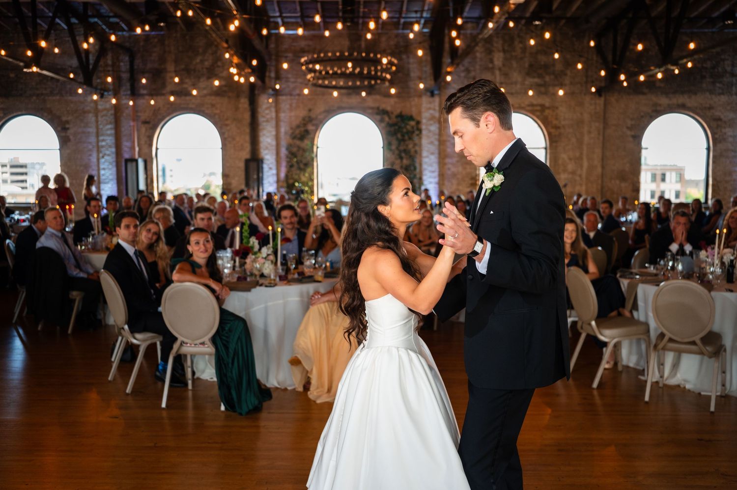 Bride and groom share their first dance at elegant wedding reception with string lights and arched windows.