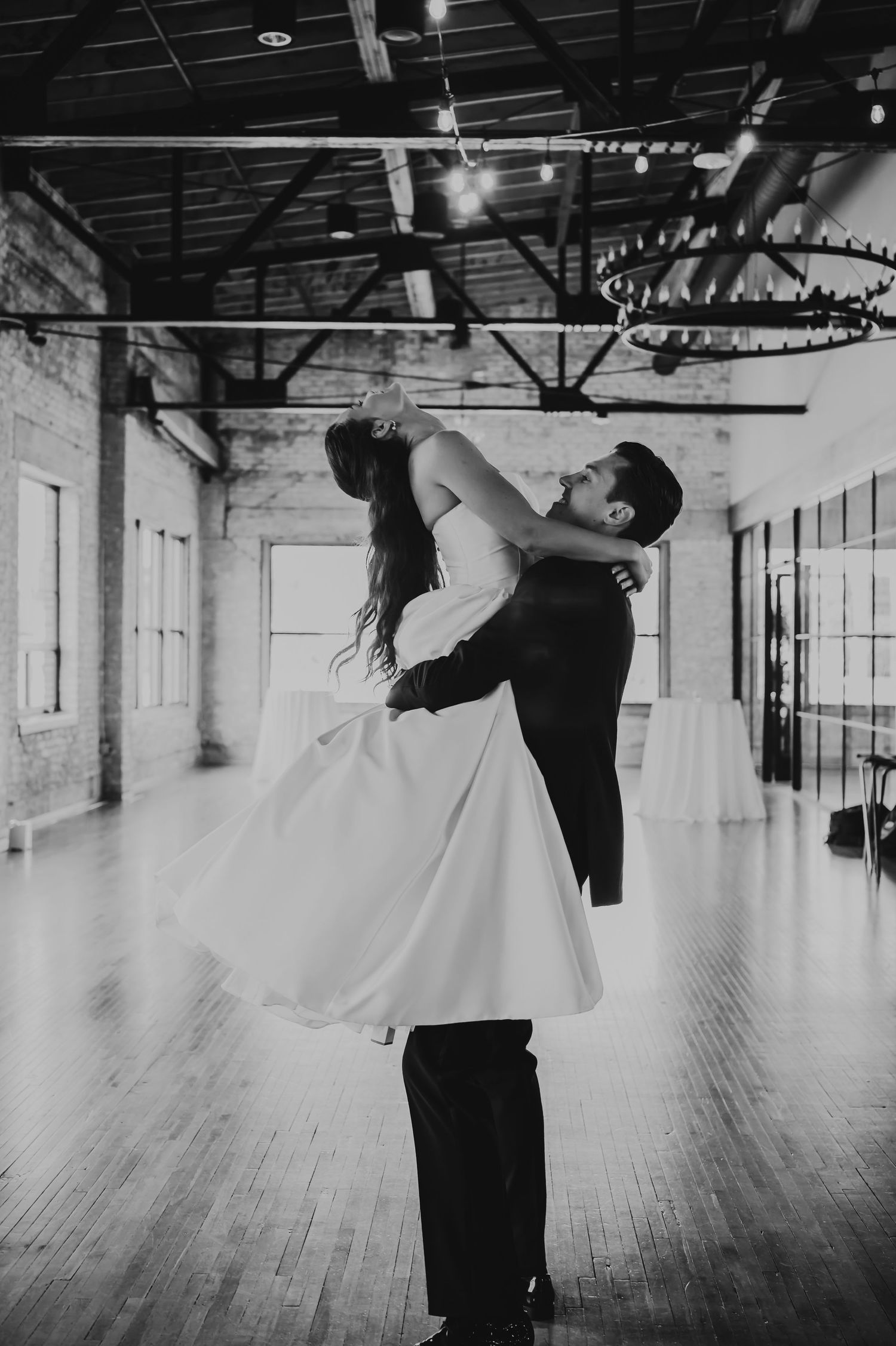 Black and white photo of groom lifting bride during first dance in elegant rustic wedding venue.