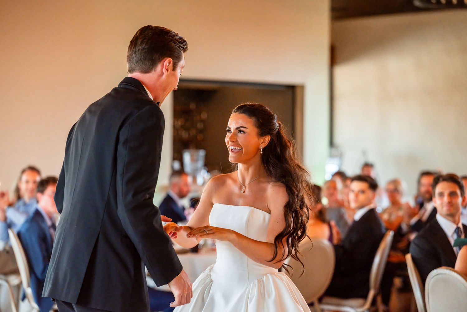 Bride smiling joyfully at groom during wedding ceremony, guests seated in background.