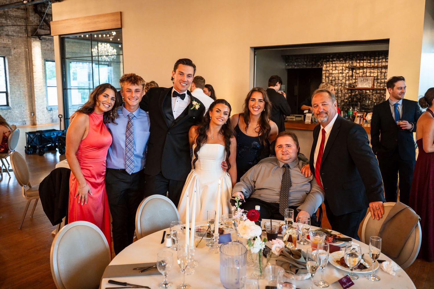 Wedding party poses together at reception table, bride in white gown with groom in tuxedo surrounded by smiling family members.